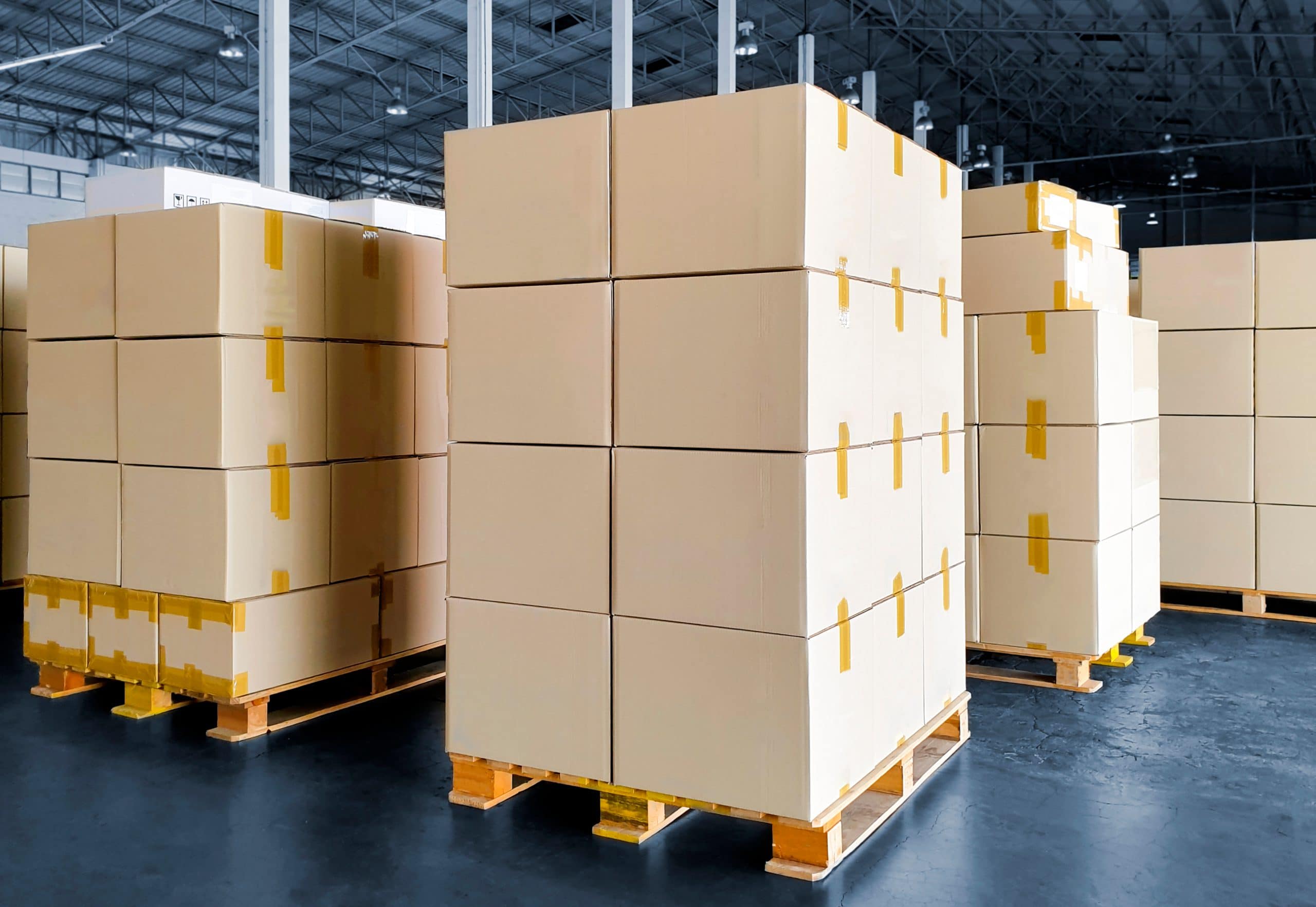 Meerdere stapels kartonnen dozen op houten pallets met open dek, omwikkeld met tape, in een magazijn.